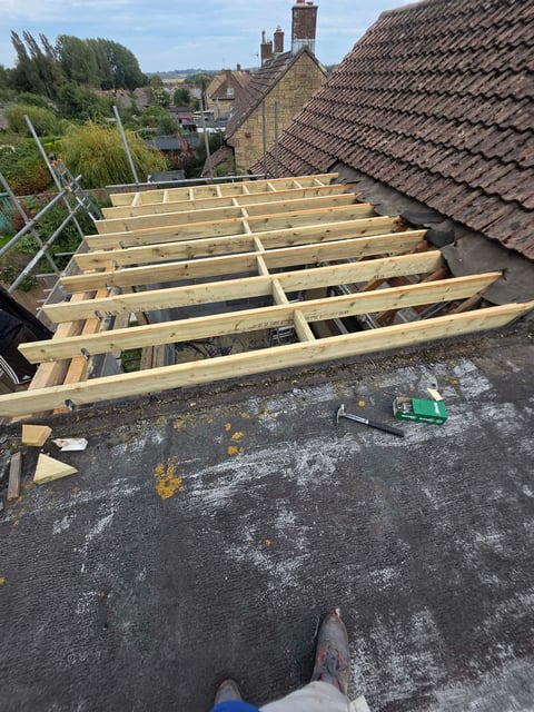 Roof framing under construction showing wooden joists and scaffolding, with rural village houses visible in background