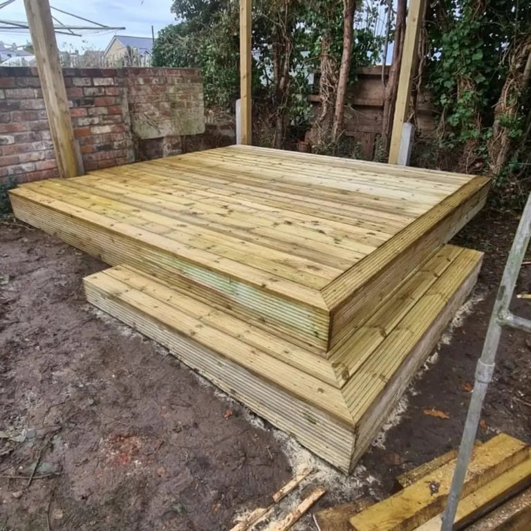 Wooden deck platform with multiple stepped levels under construction in a backyard garden area surrounded by brick walls and timber support posts