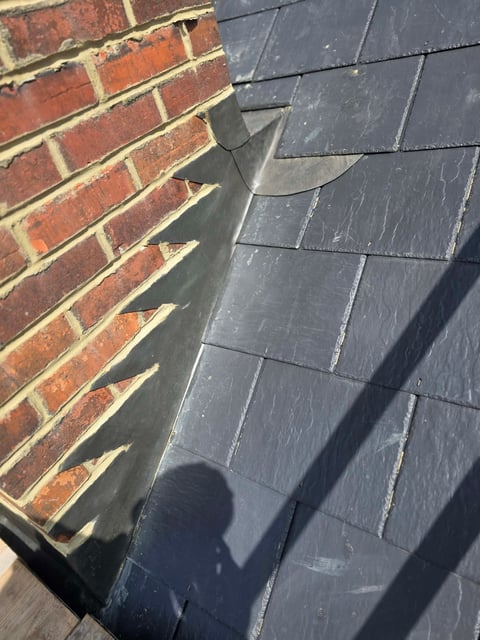Close-up of roof corner showing red brick wall meeting dark slate roof tiles with metal flashing and shadows
