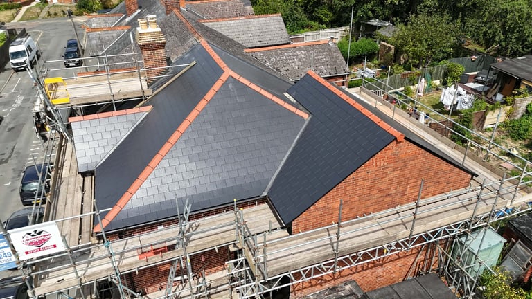 Aerial view of a roof under construction with dark tiles and orange brick sections, surrounded by scaffolding and residential buildings