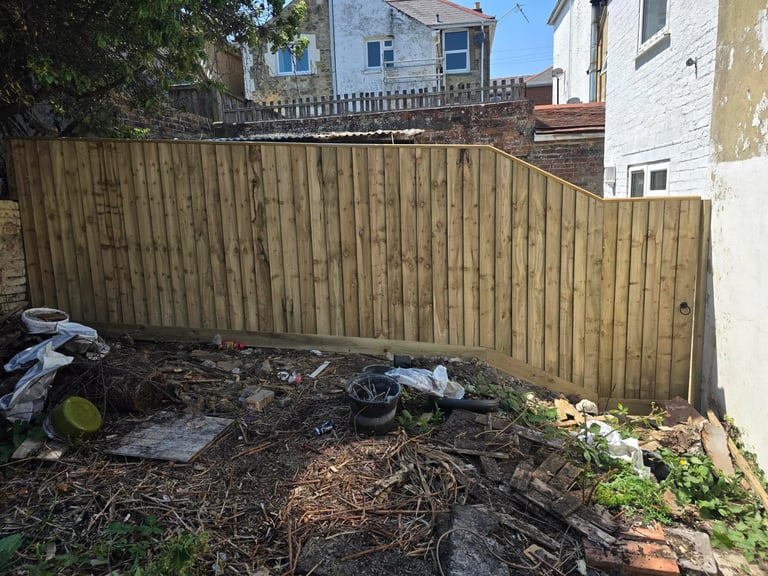Weathered wooden fence enclosing a backyard with scattered debris, logs, and green plants against residential buildings