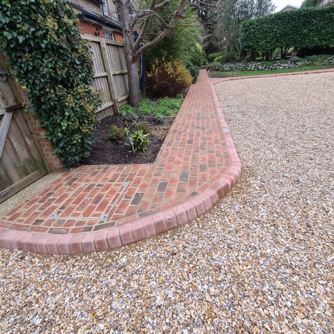 Curved red brick pathway bordering a gravel driveway with ivy-covered wooden fence