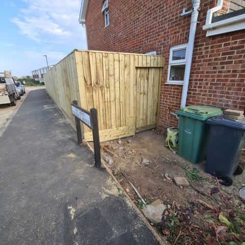Wooden gate enclosure attached to red brick house with bins nearby