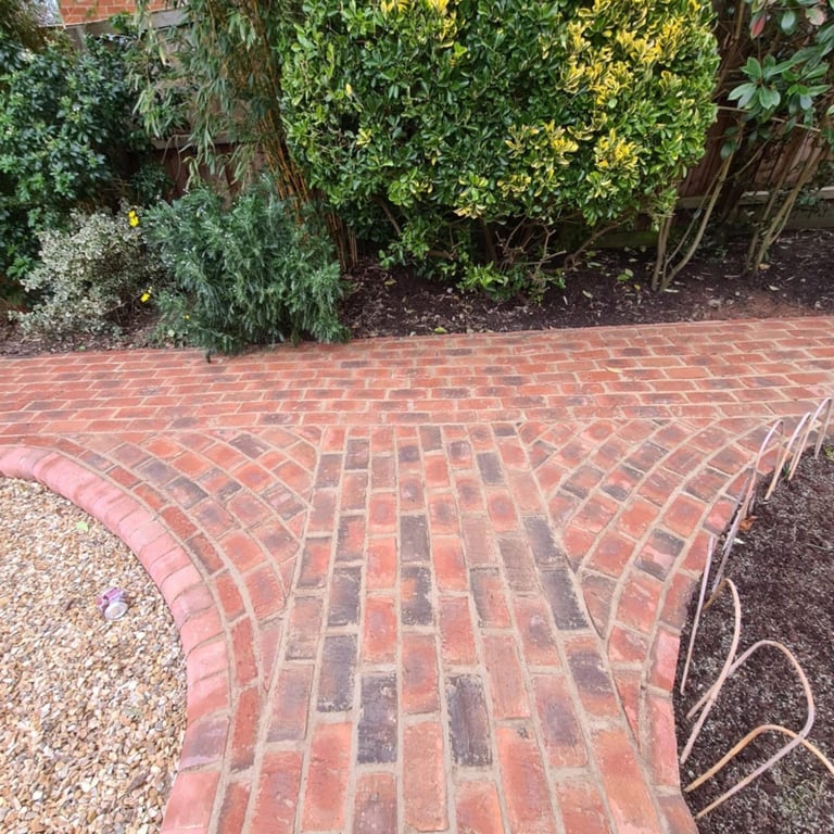 Red brick patio with herringbone pattern bordered by green shrubs and flowering plants