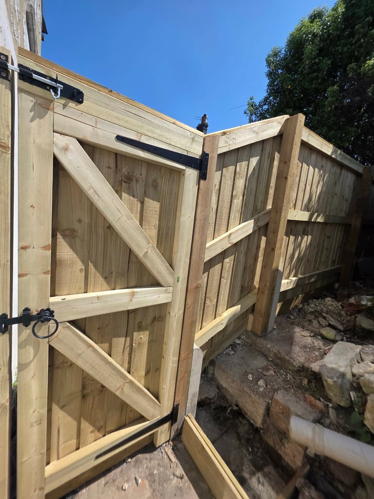 Wooden fence with open gate panels and metal hinges, clear sky and green foliage visible in background